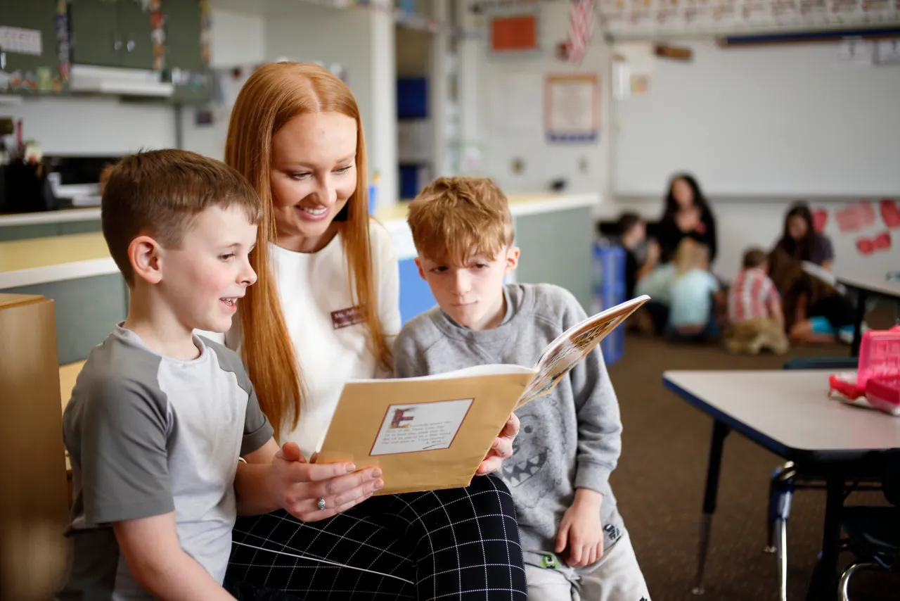 Smiling teacher reading to two young boys in a classroom overlayed with a blue icon featuring the state of Indiana and the text Service to the State